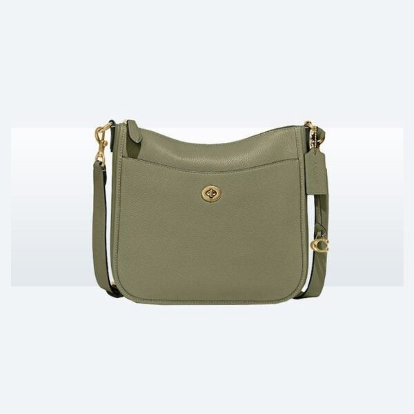 Olive green leather crossbody bag resting on an open suitcase in a sunlit hotel room.