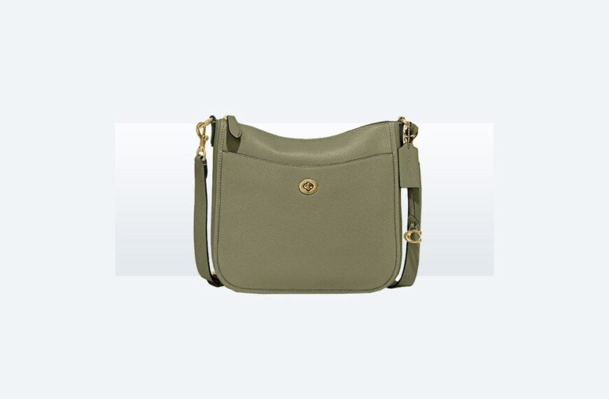 Olive green leather crossbody bag resting on an open suitcase in a sunlit hotel room.