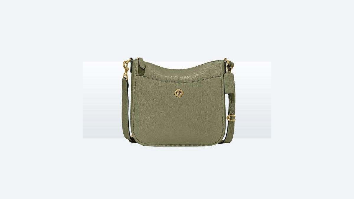 Olive green leather crossbody bag resting on an open suitcase in a sunlit hotel room.
