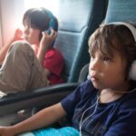A toddler wearing comfortable blue headphones while looking out an airplane window.