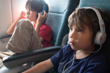 A toddler wearing comfortable blue headphones while looking out an airplane window.