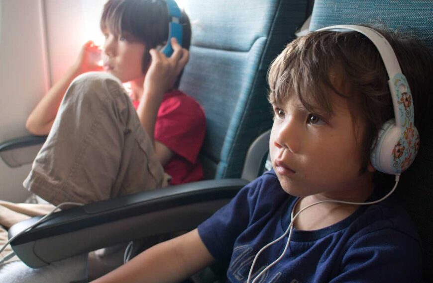 A toddler wearing comfortable blue headphones while looking out an airplane window.